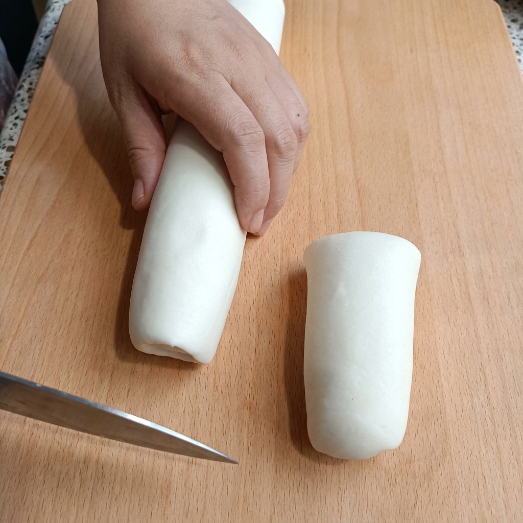 Eine Hand hält einen Zylinder aus weißem Teig auf einem Holzbrett und schneidet ihn mit einem Messer.