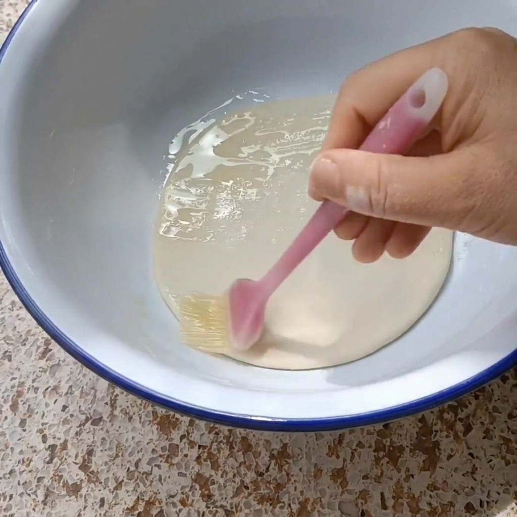Eine Hand bestreicht eine runde Teigscheibe mit einem Silikonpinsel mit Pflanzenöl.