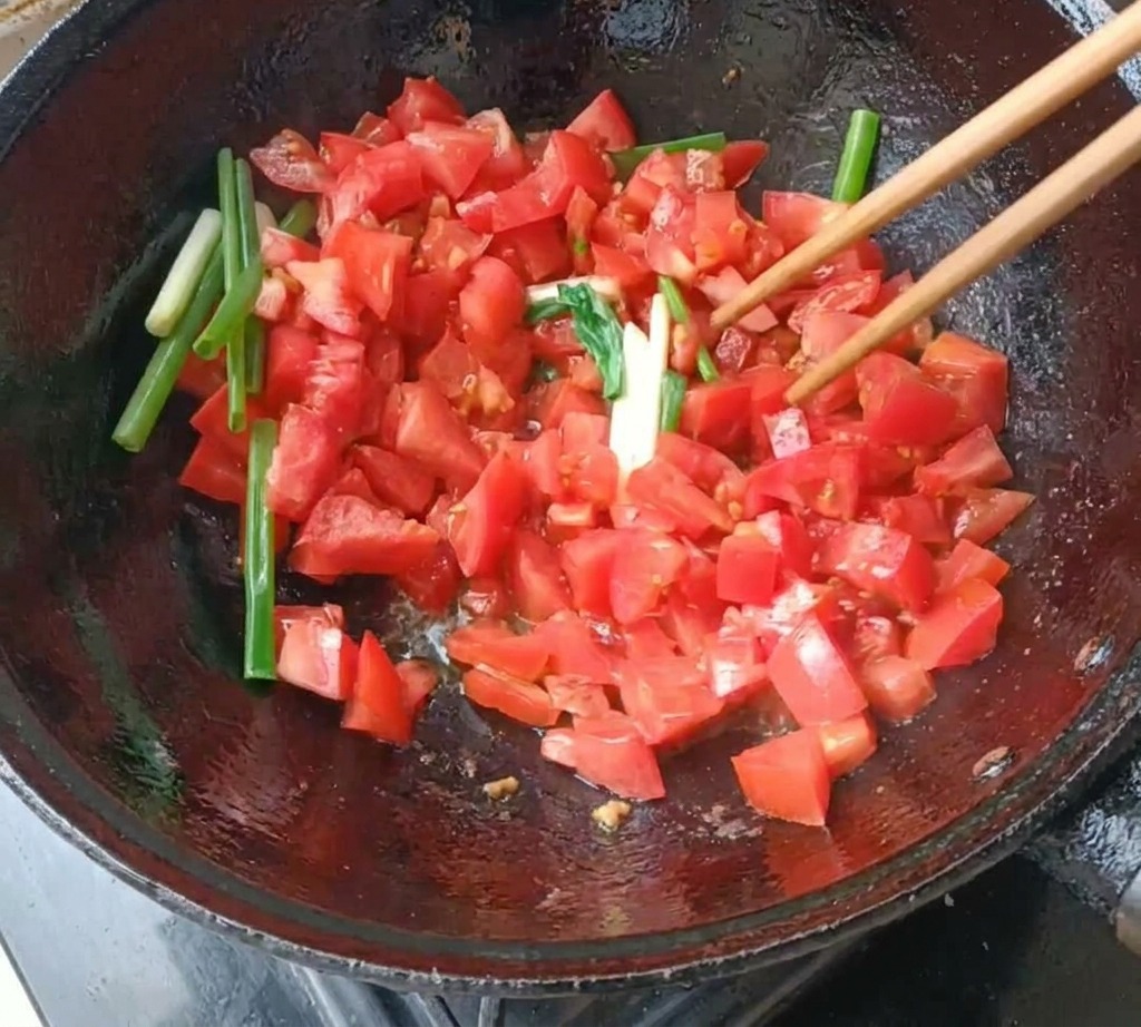 Gewürfelte Tomaten und Frühlingszwiebelstücke werden in einem dunklen Wok mit Stäbchen angebraten.