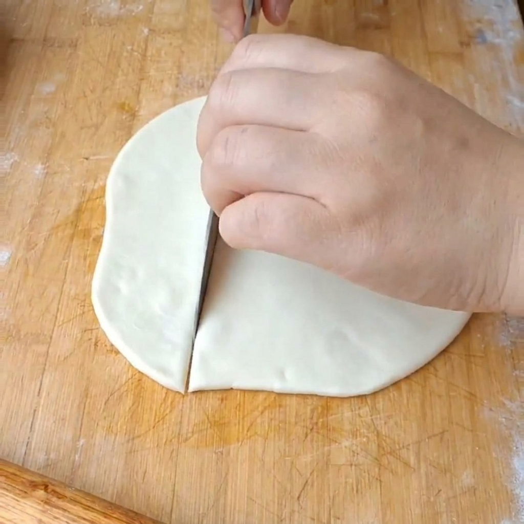 Eine Hand schneidet eine flache Teigscheibe mit einem großen Messer in breite Streifen.