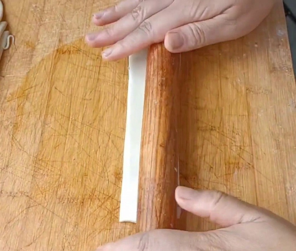 Nahaufnahme von Händen, die einen langen Teigstreifen mit einem Nudelholz flach rollen.
