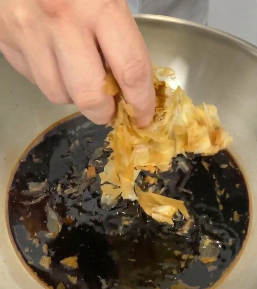 Eine Hand gibt helle, hauchdünne getrocknete Bonitoflocken in die dunkle Flüssigkeit im Topf.