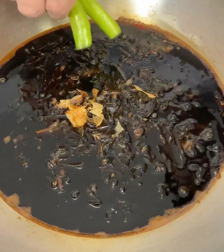 Eine Hand legt zwei leuchtend grüne Chilischoten in den Topf mit der dunklen Sauce und den Bonitoflocken.