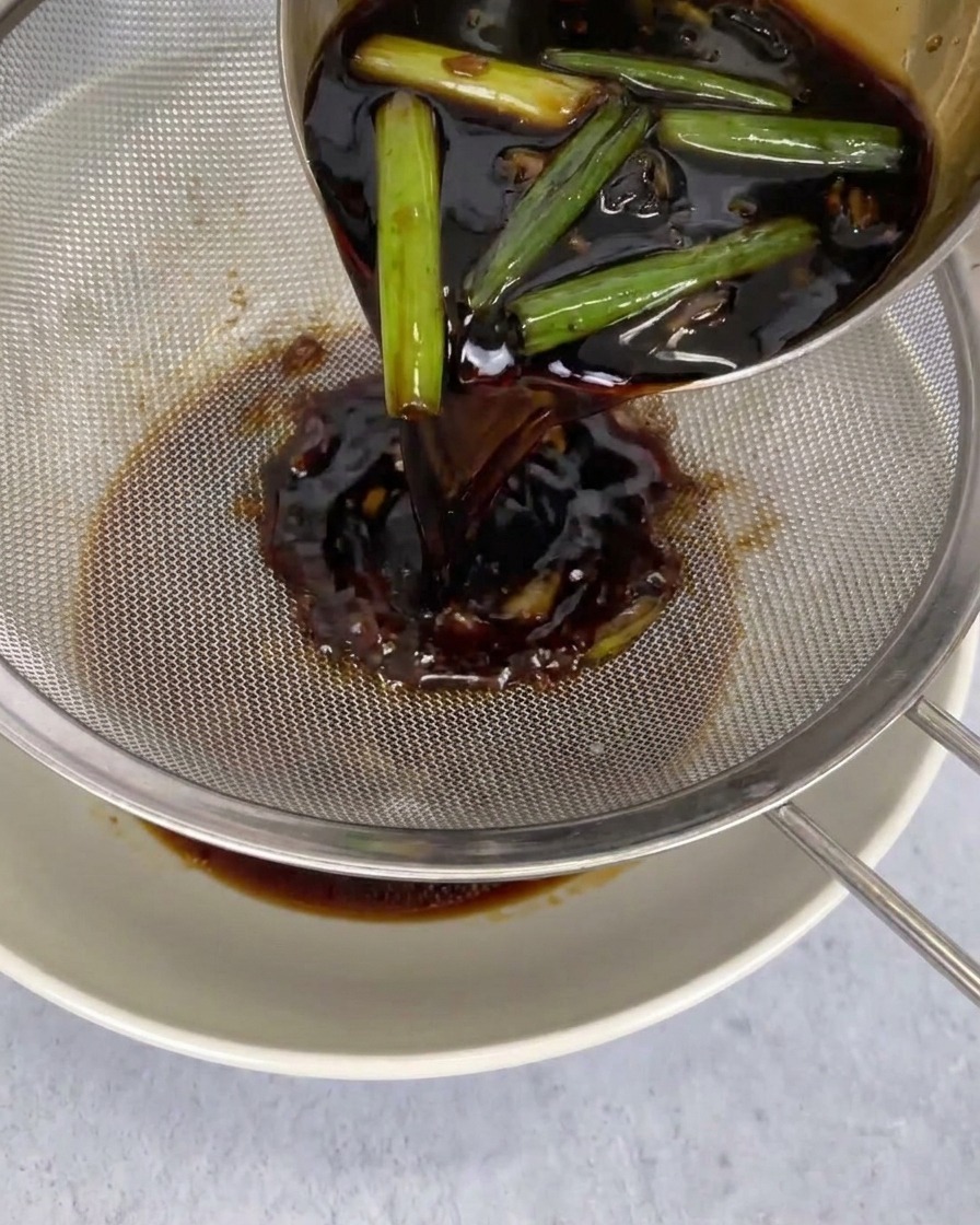 Eine dunkle Sauce wird mit einer Kelle durch ein Metallsieb in eine weiße Schüssel gegossen, wobei Chilis und Flocken im Sieb bleiben.