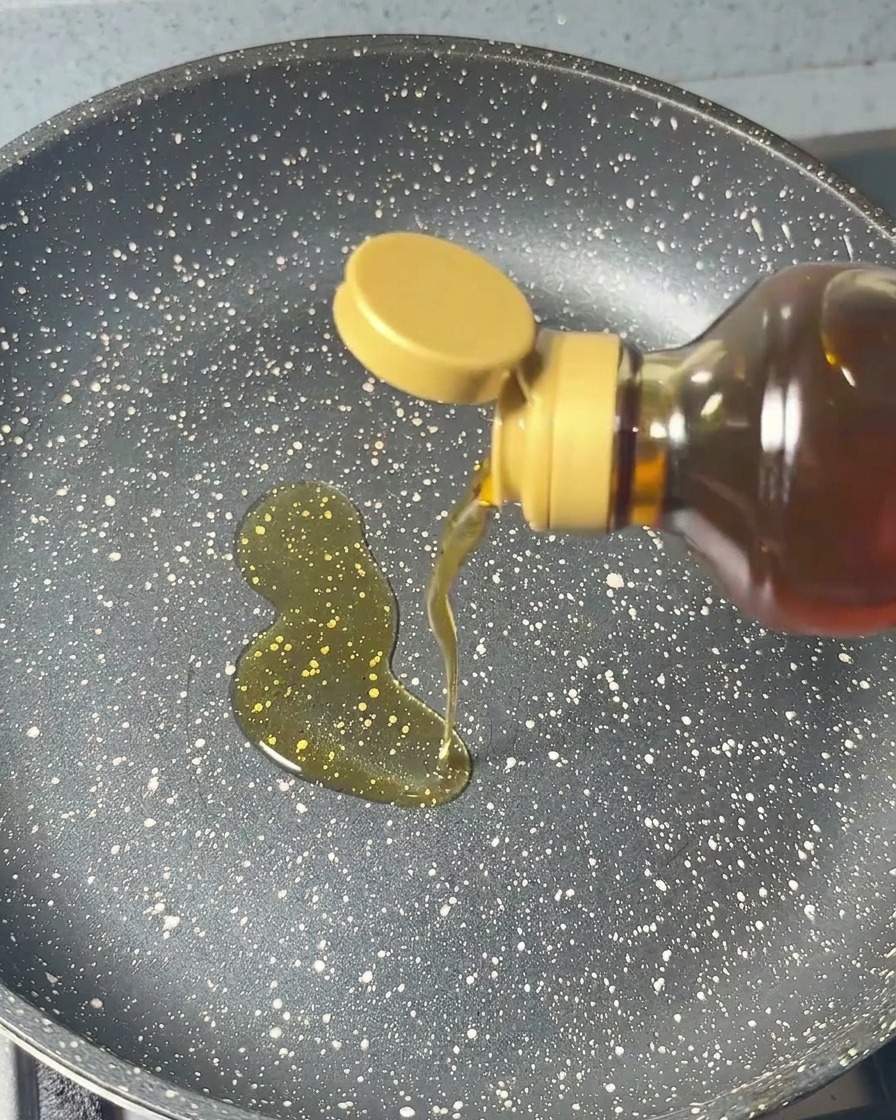 Sesamöl wird aus einer Flasche kreisförmig in eine dunkle, gesprenkelte Pfanne gegossen.