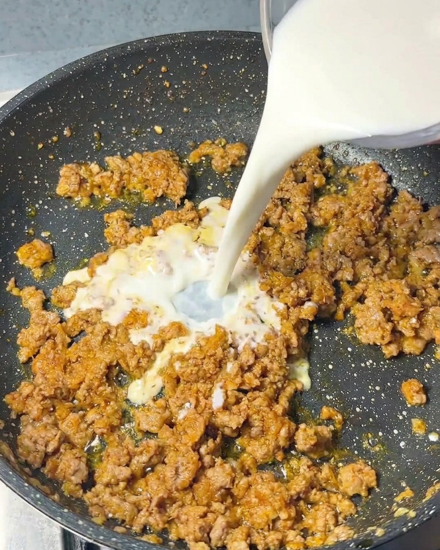 Ein Strahl weißer Milch wird aus einem Glasgefäß in die Pfanne mit dem Fleisch gegossen.