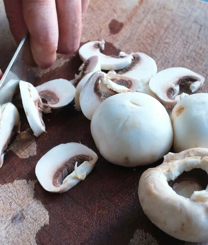 Nahaufnahme eines Kochmessers, das weiße Champignons auf einem Holzschneidebrett in Scheiben schneidet.