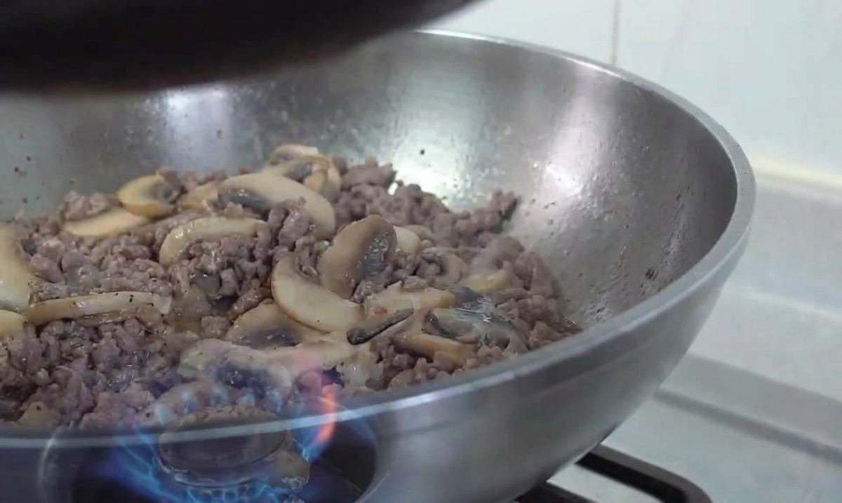 Ein Edelstahl-Wok mit Fleisch und Pilzen wird über einer Gasflamme geschwenkt.
