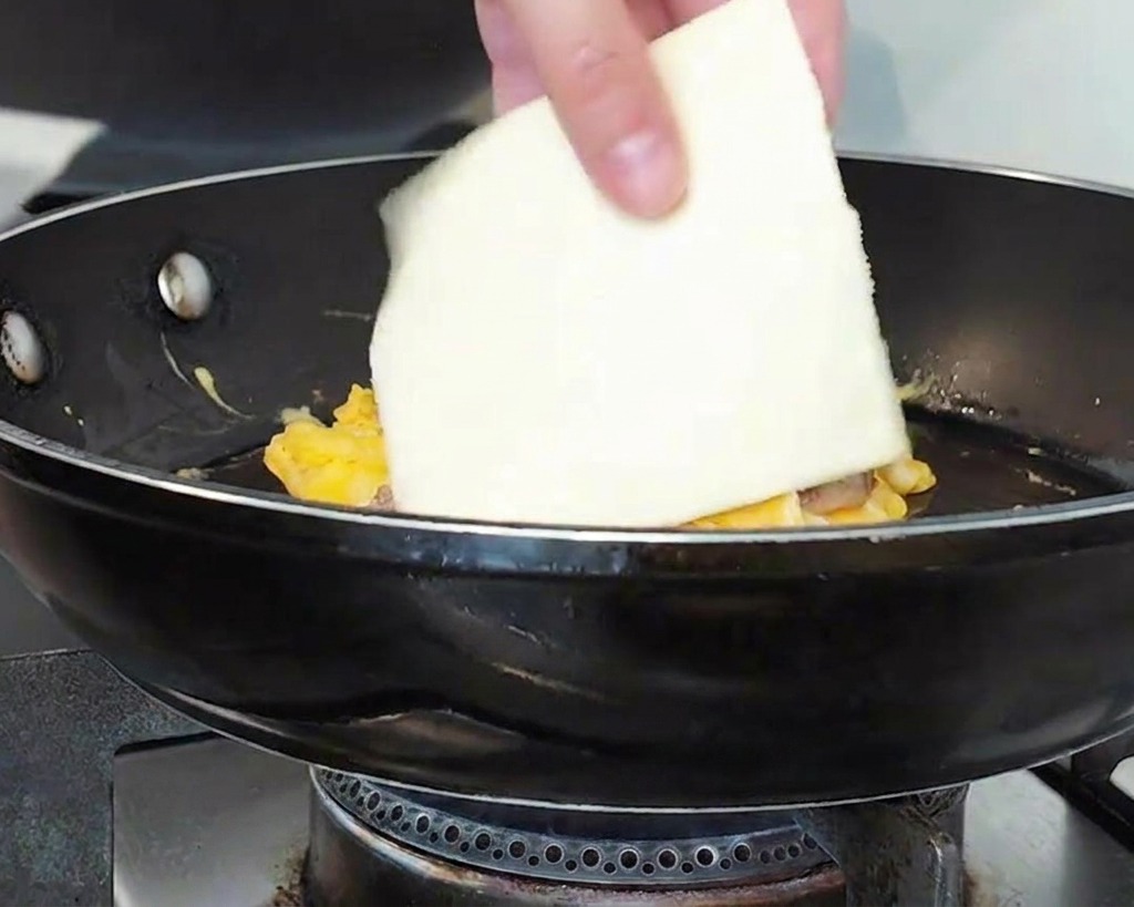 Eine Hand legt eine Scheibe Käse auf den Fleisch-Ei-Stapel in der Pfanne.