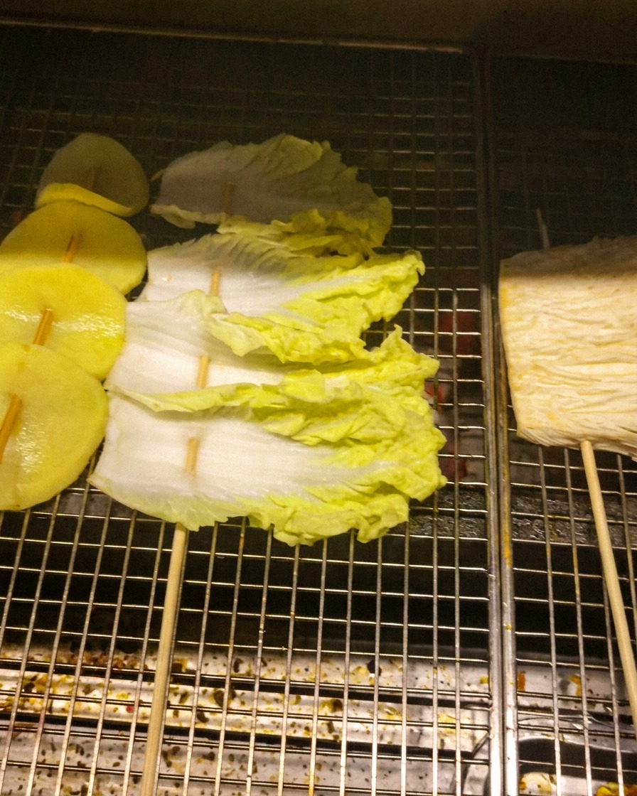 Kartoffelscheiben, Kohlblätter und Enoki-Pilze auf Spießen auf einem Grillrost über Kohlen.