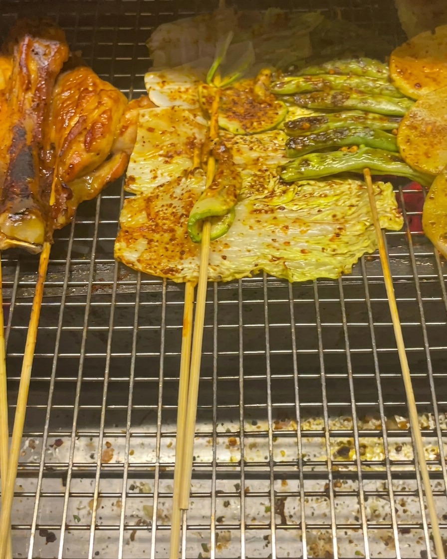 Hähnchenschenkel, Baby-Kohl, Zucchini, grüne Bohnen und Kartoffelscheiben auf einem Grillrost.