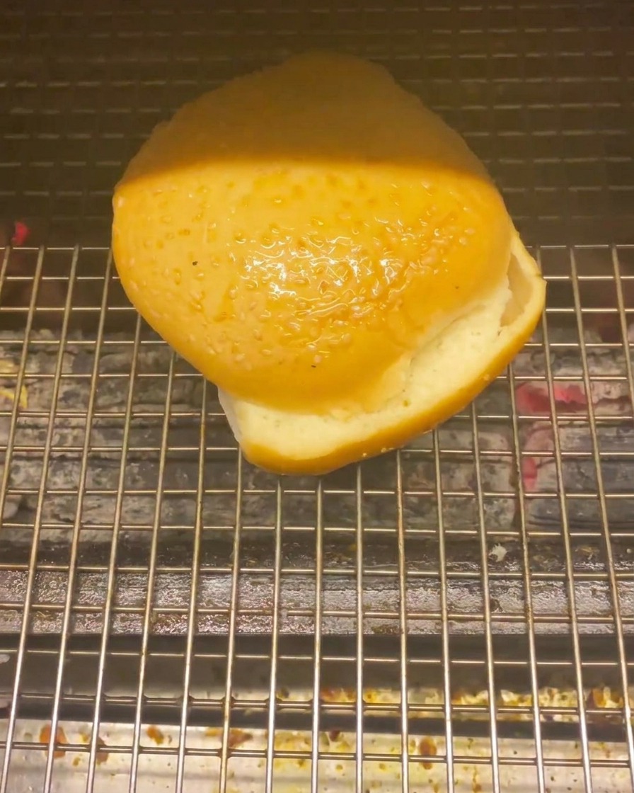 Ein Burgerbrötchen auf einem Grillrost über heißer Kohle zum Rösten.