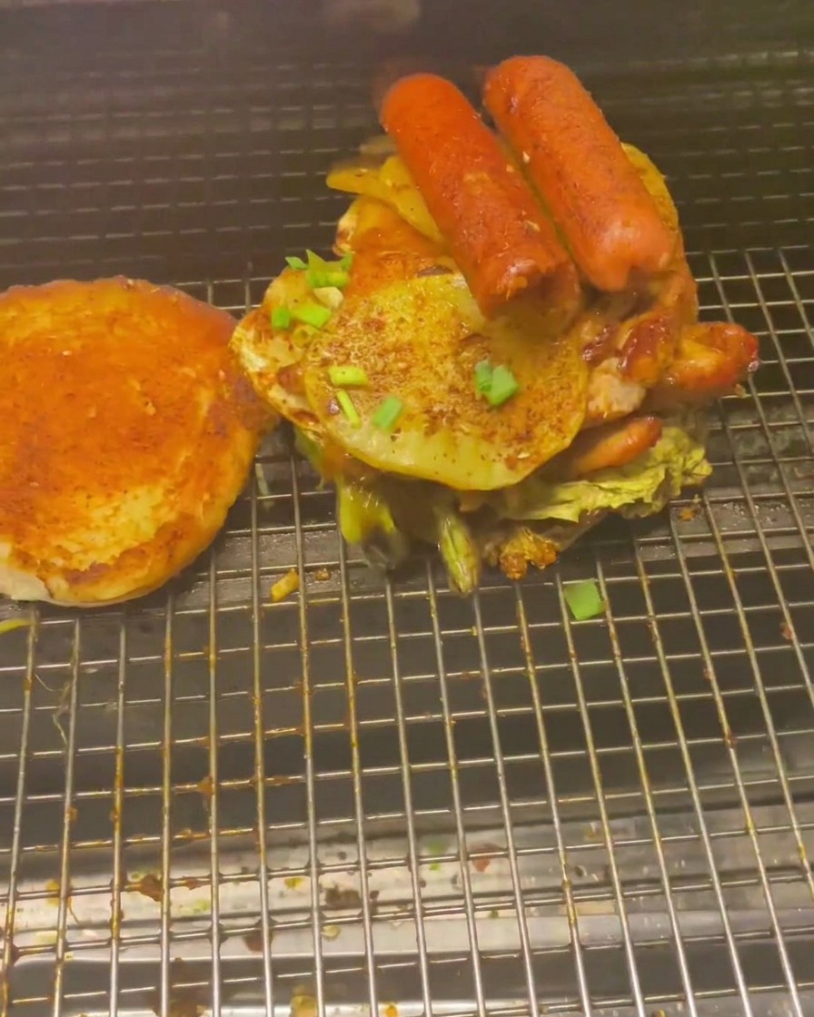 Ein hoher Stapel aus Grillfleisch, Würstchen und Kartoffelscheiben auf einem Burgerbrötchen.