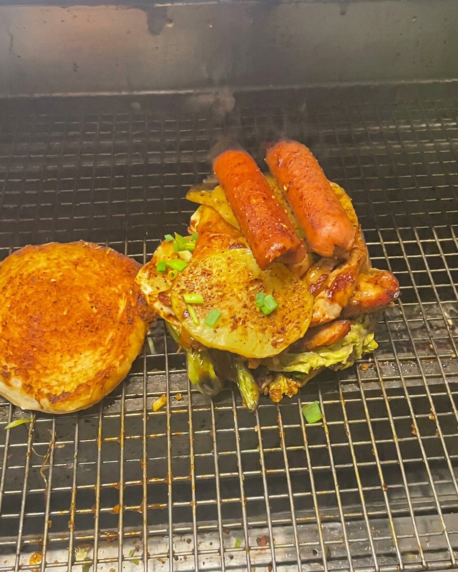 Ein riesiger Fleisch-Gemüse-Turm auf einem Brötchen, die obere Brötchenhälfte liegt daneben auf dem Grill.