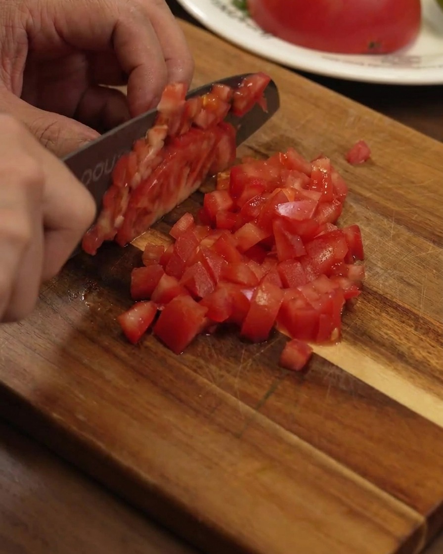 Eine reife rote Tomate wird auf einem Holzbrett mit einem Kochmesser in kleine, gleichmäßige Würfel geschnitten.