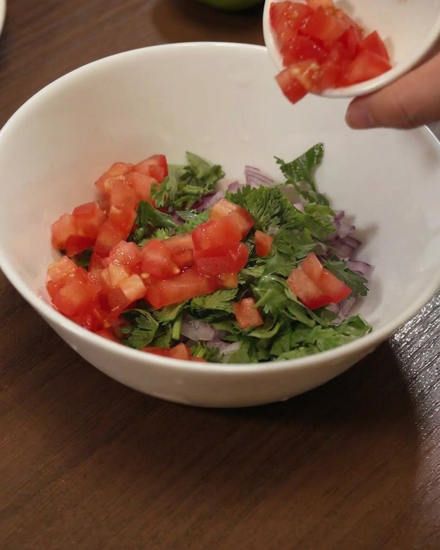 Gewürfelte rote Tomaten werden aus einer Schale in eine weiße Schüssel zu Koriander und roten Zwiebeln gegeben.