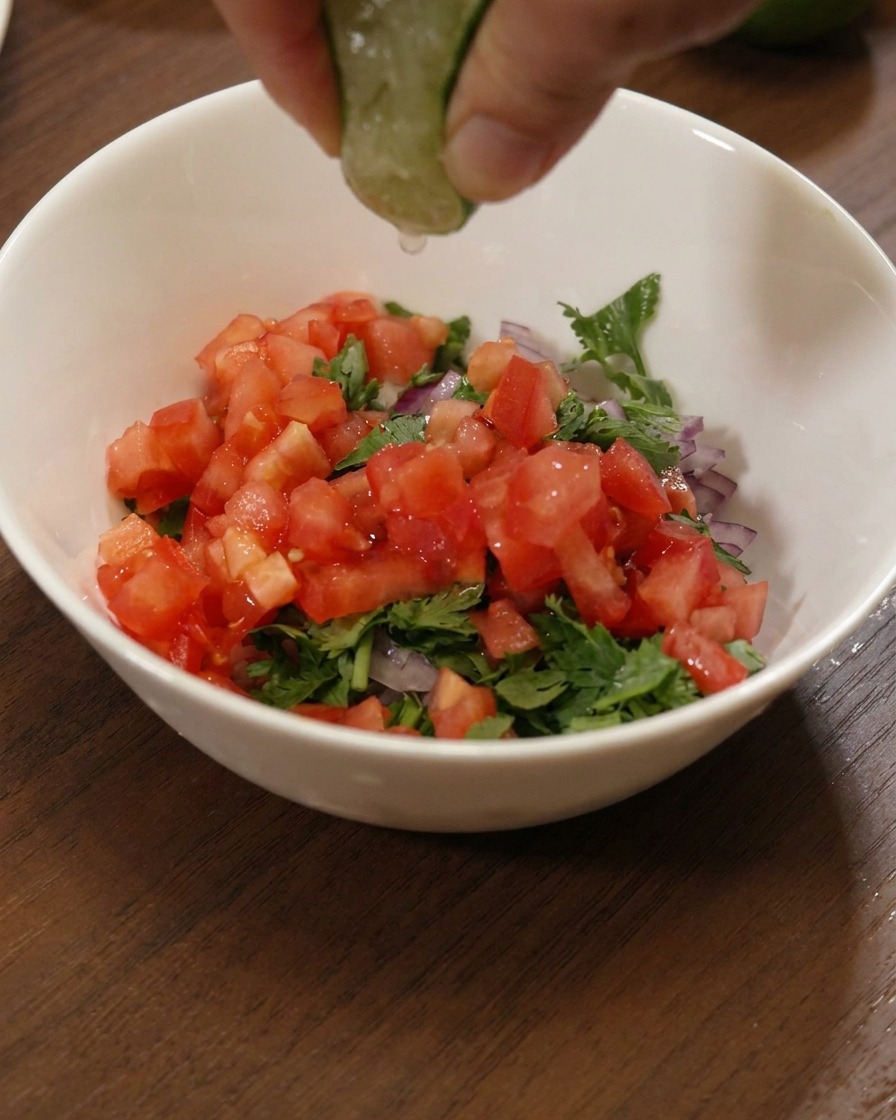 Eine Hand presst eine frische Limettenspalte über eine Schüssel mit Tomaten, Zwiebeln und Koriander.