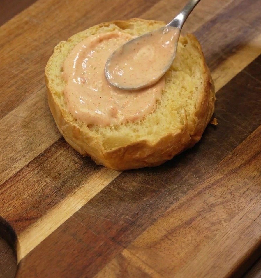 Ein silberner Löffel verstreicht eine dicke Schicht orangefarbene Sauce auf der Innenseite eines Brioche-Brötchens.