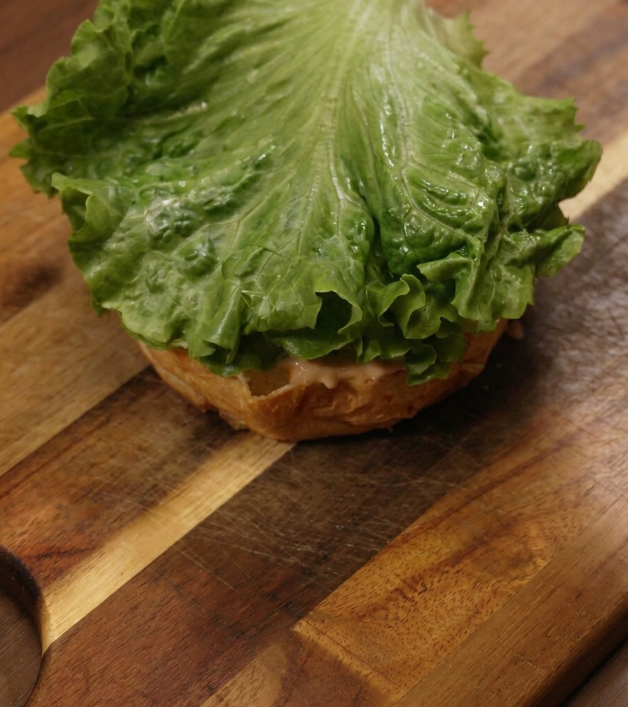 Ein knackiges grünes Salatblatt liegt auf der bestrichenen Unterseite eines Burger-Brötchens.