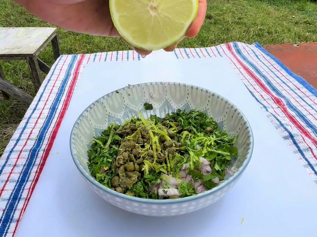 Eine Hand presst eine halbe frische Limette über einer gemusterten Schüssel mit gehackten Kräutern, Kapern und Schalotten aus.