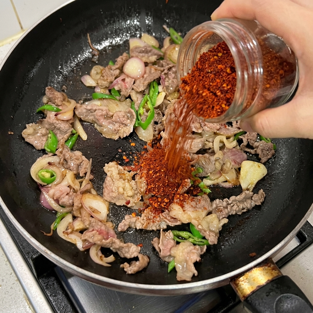 Rotes Chilipulver wird aus einem Glas über Rindfleisch, Zwiebeln und grüne Chilis in einer schwarzen Pfanne gestreut.