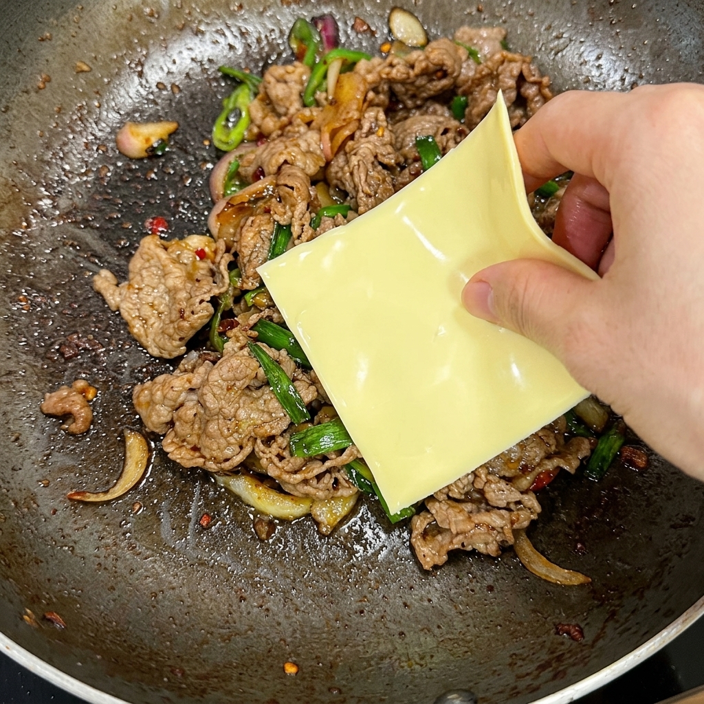 Eine Hand legt eine quadratische Scheibe gelben Käse auf die heiße Rindfleisch-Mischung in einer Pfanne.