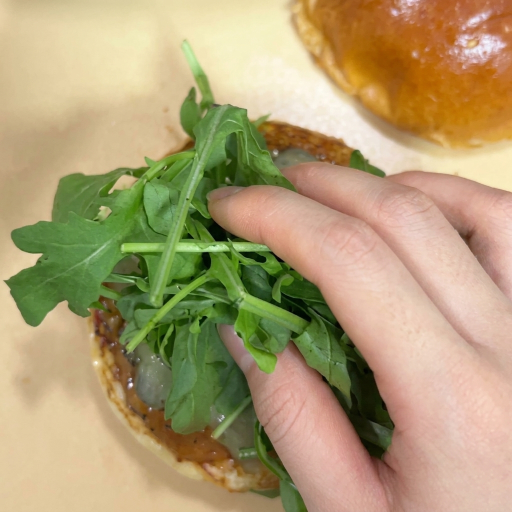 Eine Hand legt eine frische Portion grünen Rucola auf die Gurkenscheiben einer Brötchenhälfte.
