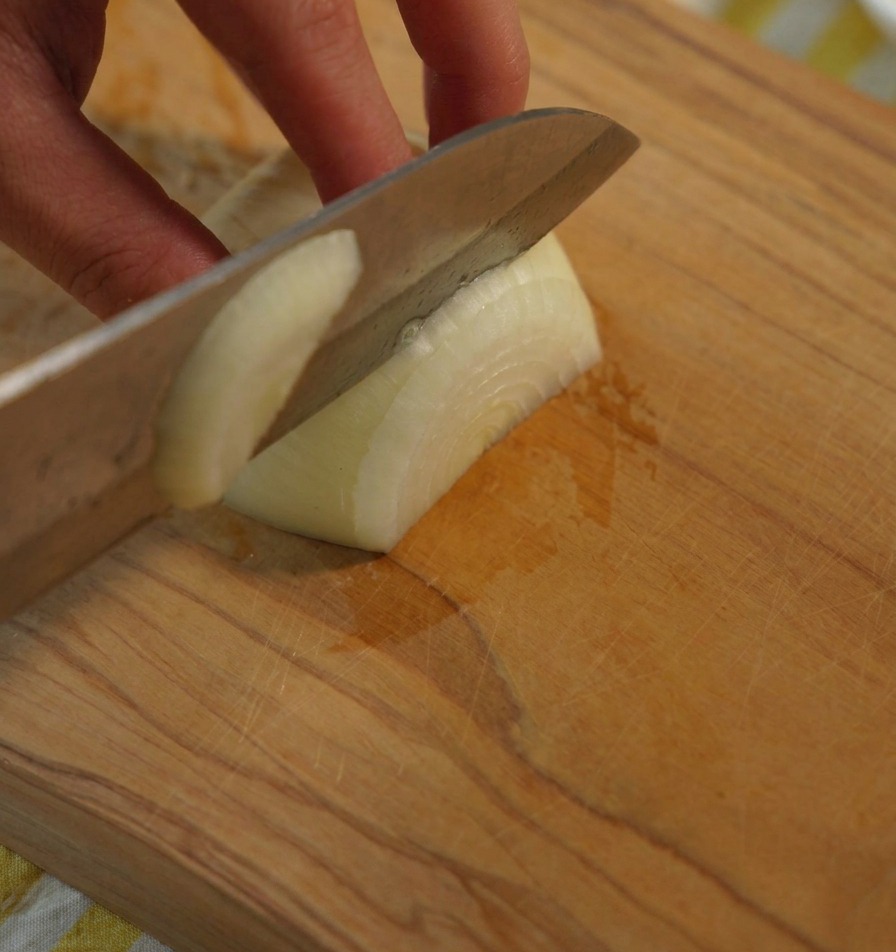 Eine Nahaufnahme einer Hand, die eine weiße Zwiebel hält, während ein scharfes Kochmesser sie auf einem Holzbrett in gleichmäßige Stücke schneidet.