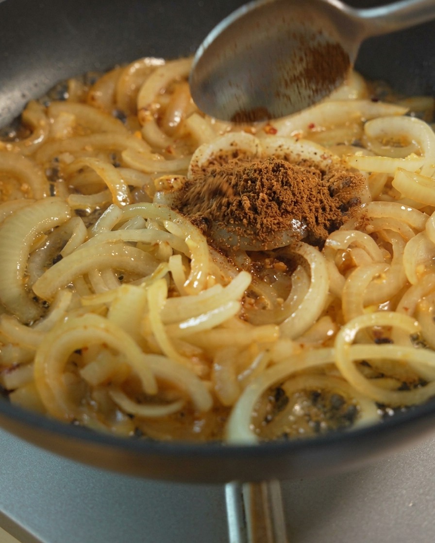 Zwiebelscheiben werden in einer Pfanne angedünstet, während ein Löffel braunen Zucker und flüssige Gewürze zum Karamellisieren hinzufügt.