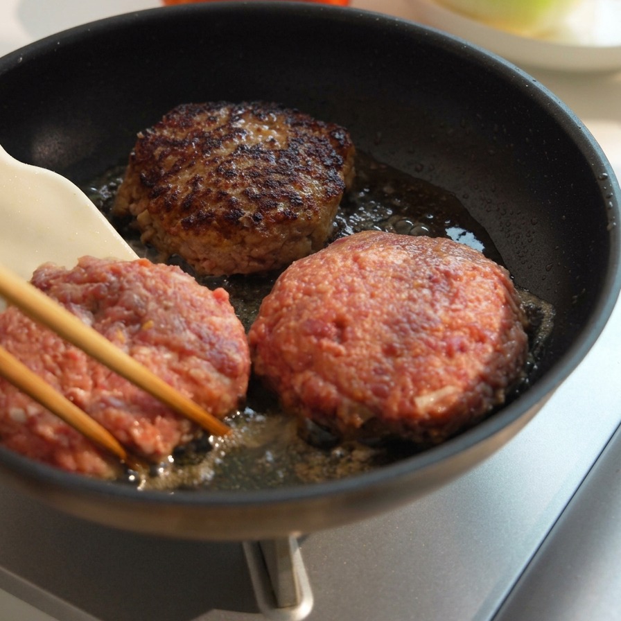 Drei dicke Rinder-Patties braten in einer Pfanne, wobei eines umgedreht wurde, um eine kräftig gebräunte, karamellisierte Kruste zu zeigen.