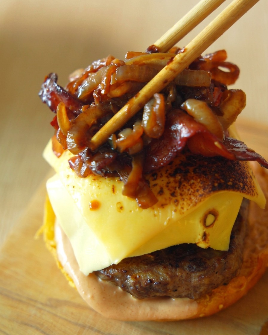 Knusprige Speckstreifen und süße karamellisierte Zwiebeln werden mit Holzstäbchen vorsichtig auf ein mit Käse bedecktes Burger-Patty gelegt.