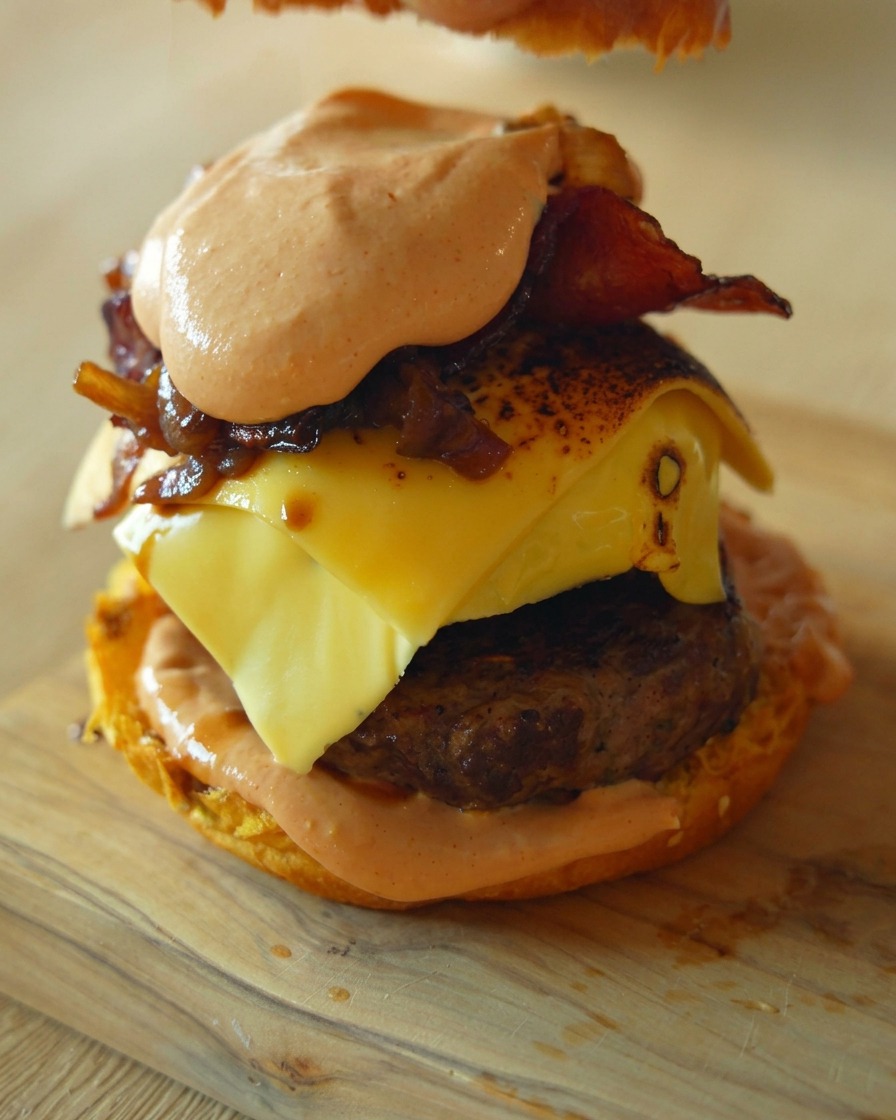 Eine Nahaufnahme, die zeigt, wie eine Hand das obere Brötchen auf einen hohen Gourmet-Burger setzt, der mit Patty, geschmolzenem Käse und Zwiebeln geschichtet ist.