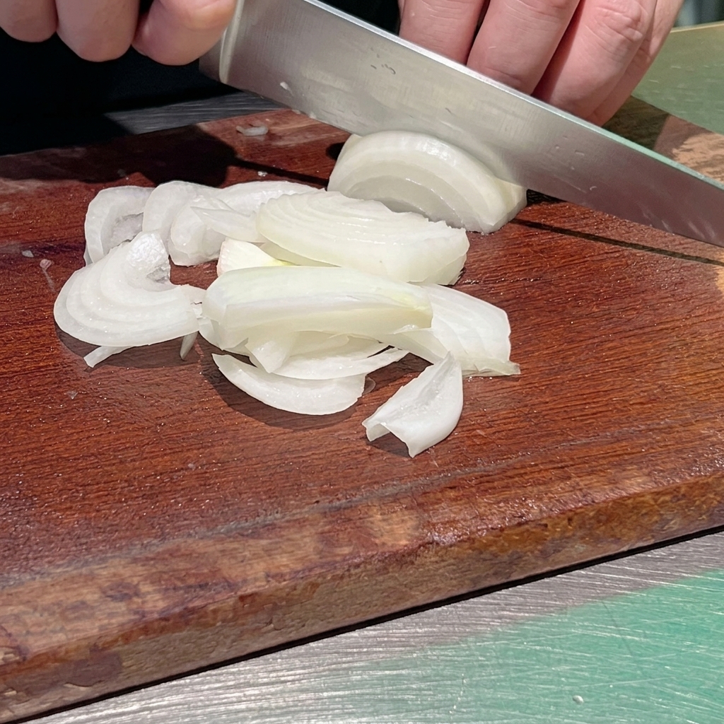 Nahaufnahme eines Kochs, der eine weiße Zwiebel mit einem scharfen Küchenmesser auf einem dunklen Holzbrett in feine Streifen schneidet.