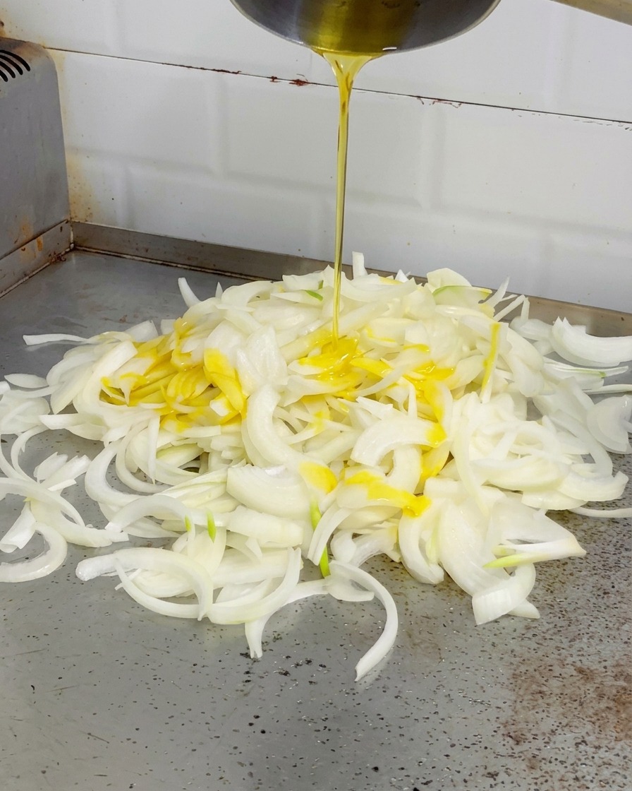 Geschmolzene gelbe Butter wird aus einem Silbertopf über einen Haufen Zwiebeln auf einer Grillplatte gegossen.