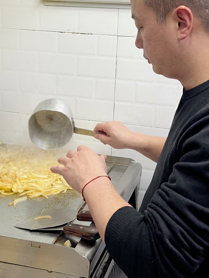 Ein Koch gießt Wasser aus einer kleinen Metallkelle auf weich werdende Zwiebeln auf einer gewerblichen Grillplatte.
