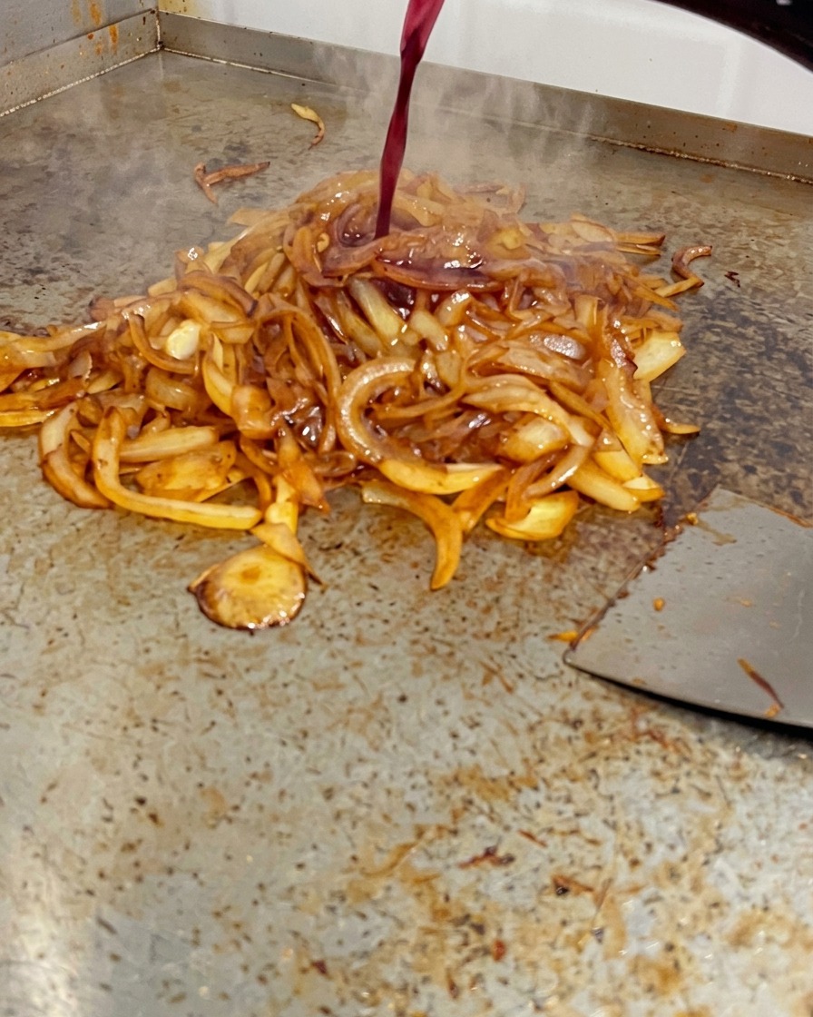Rotwein wird aus einer Flasche über tiefbraune Zwiebeln auf der heißen Grillplatte gegossen.