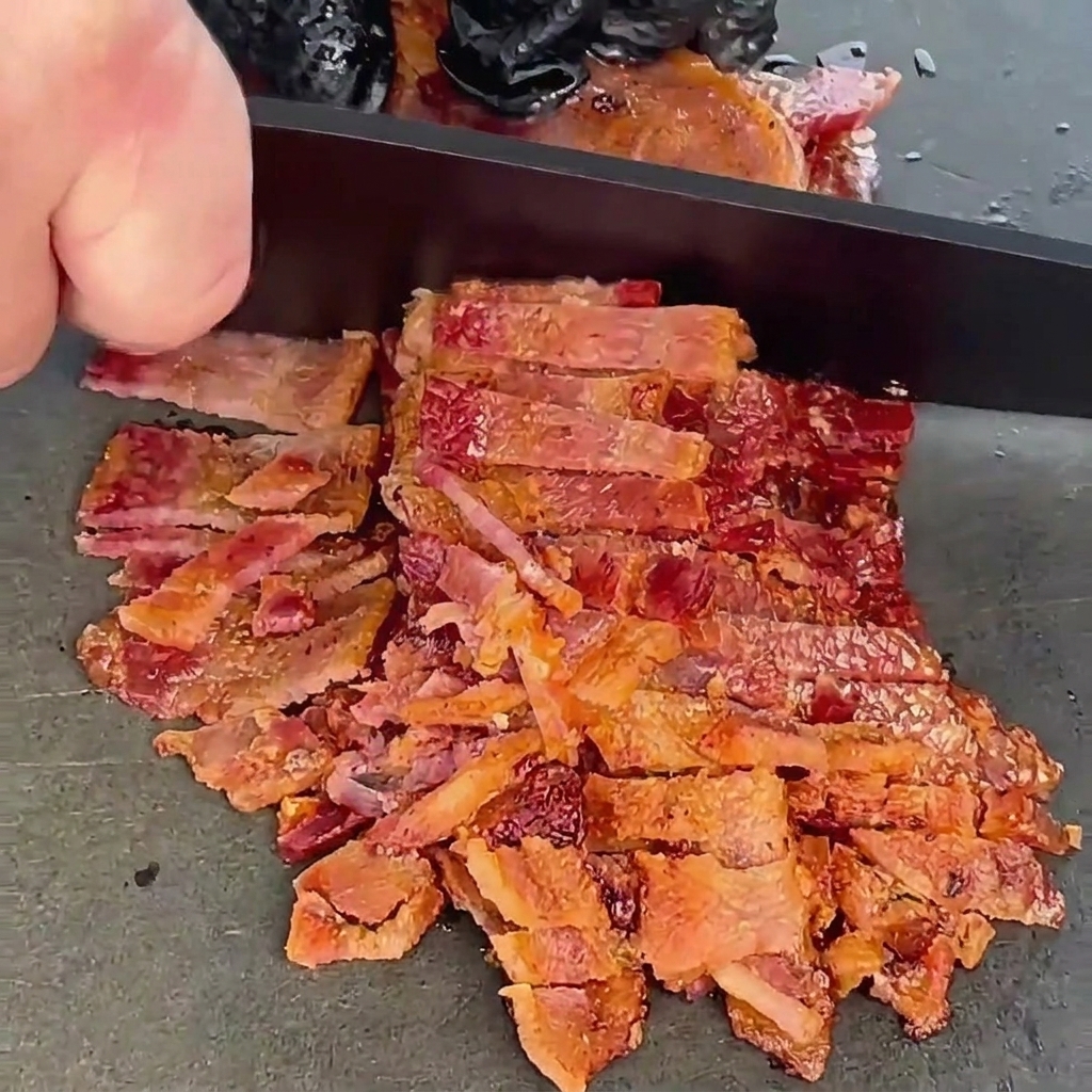 Gebratene, knusprige Baconstreifen werden mit einem schwarzen Kochmesser auf einer dunkelgrauen Arbeitsfläche fein gehackt.