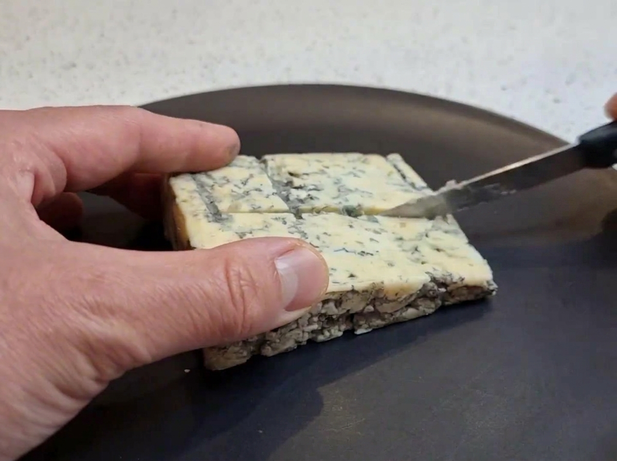 Eine Hand schneidet ein quadratisches Stück Blauschimmelkäse mit einem kleinen Messer auf einem dunklen Teller in kleinere Stücke.