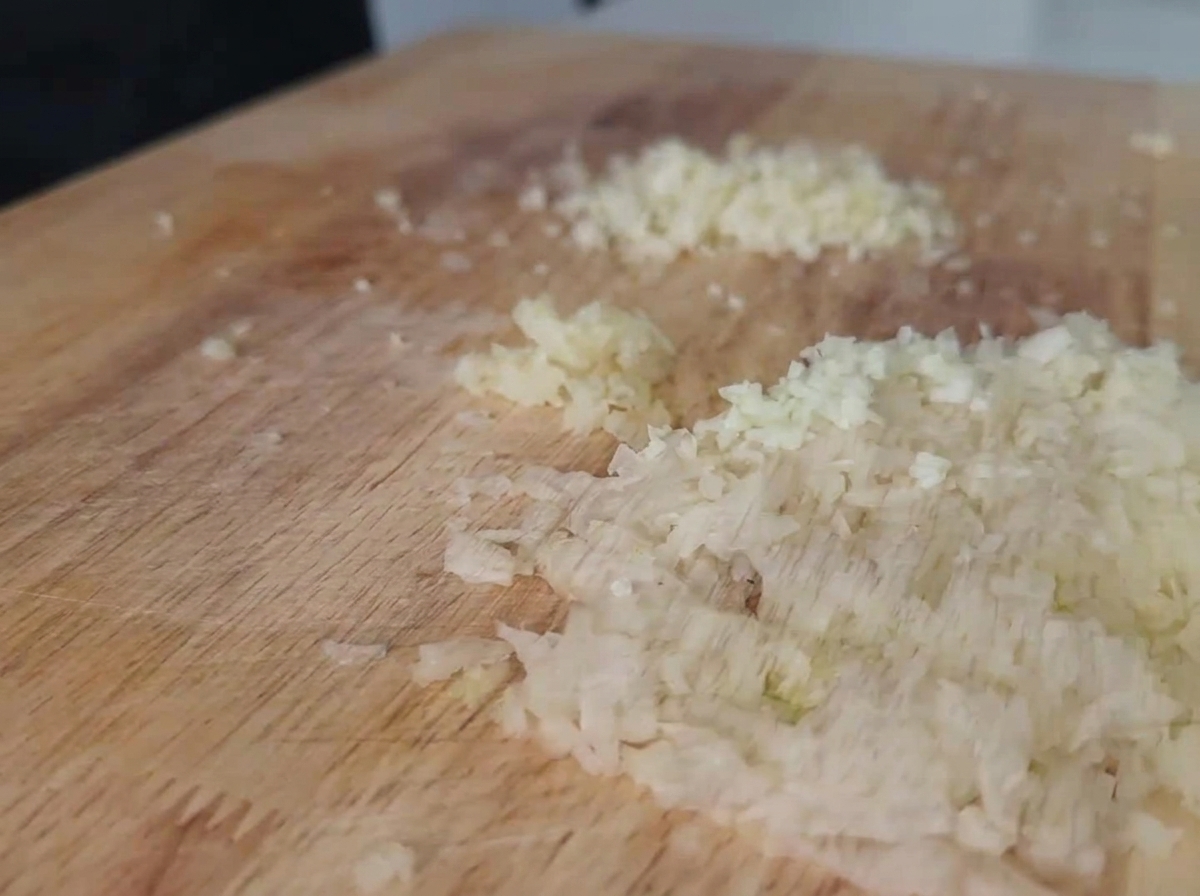 Fein gehackte weiße Zwiebeln und Knoblauch liegen verteilt auf einem hellen Holzschneidebrett.