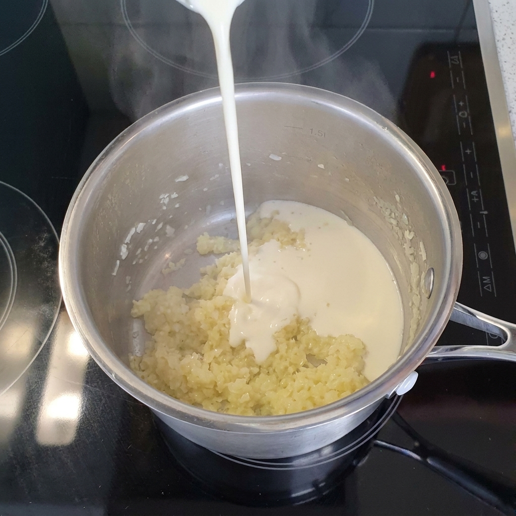 Ein Strahl flüssige Sahne wird in einen Edelstahltopf mit gedünsteten Zwiebeln und Knoblauch gegossen.