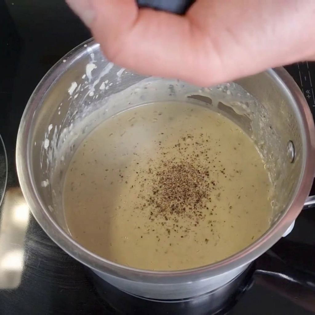 Eine Hand mahlt frischen schwarzen Pfeffer aus einer Mühle direkt in eine cremige Sauce in einem Edelstahltopf auf einem schwarzen Herd.