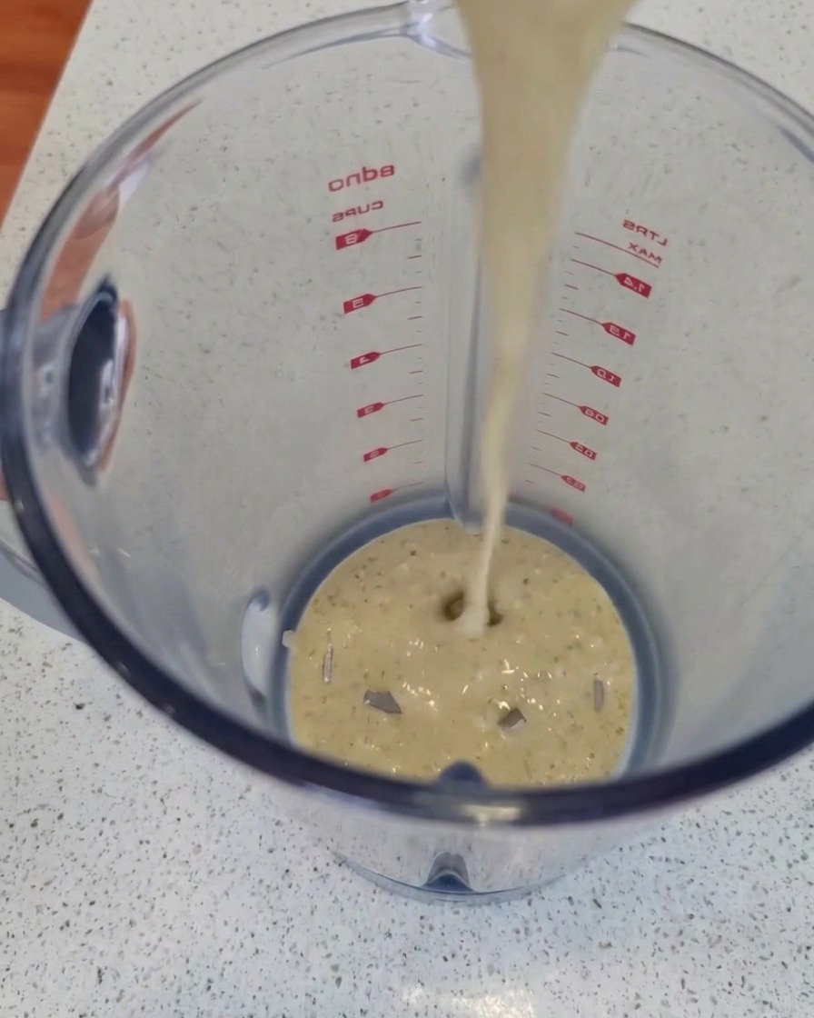 Ein Strahl cremige, fein gesprenkelte Blauschimmelkäsesauce wird von oben in einen klaren Mixerbehälter aus Kunststoff auf einer Küchenarbeitsplatte gegossen.