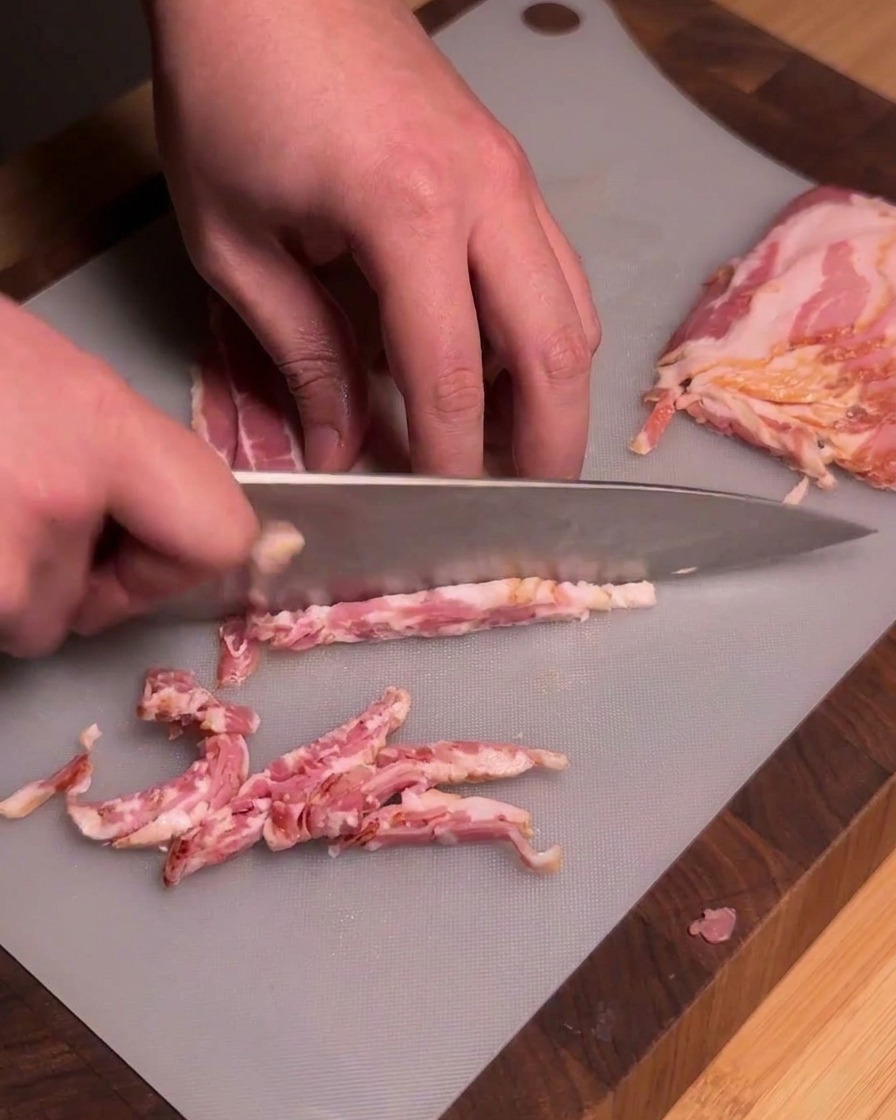 Ein Koch schneidet mit einem scharfen Küchenmesser rohe Baconstreifen auf einem grauen Schneidebrett in gleichmäßige Stücke.