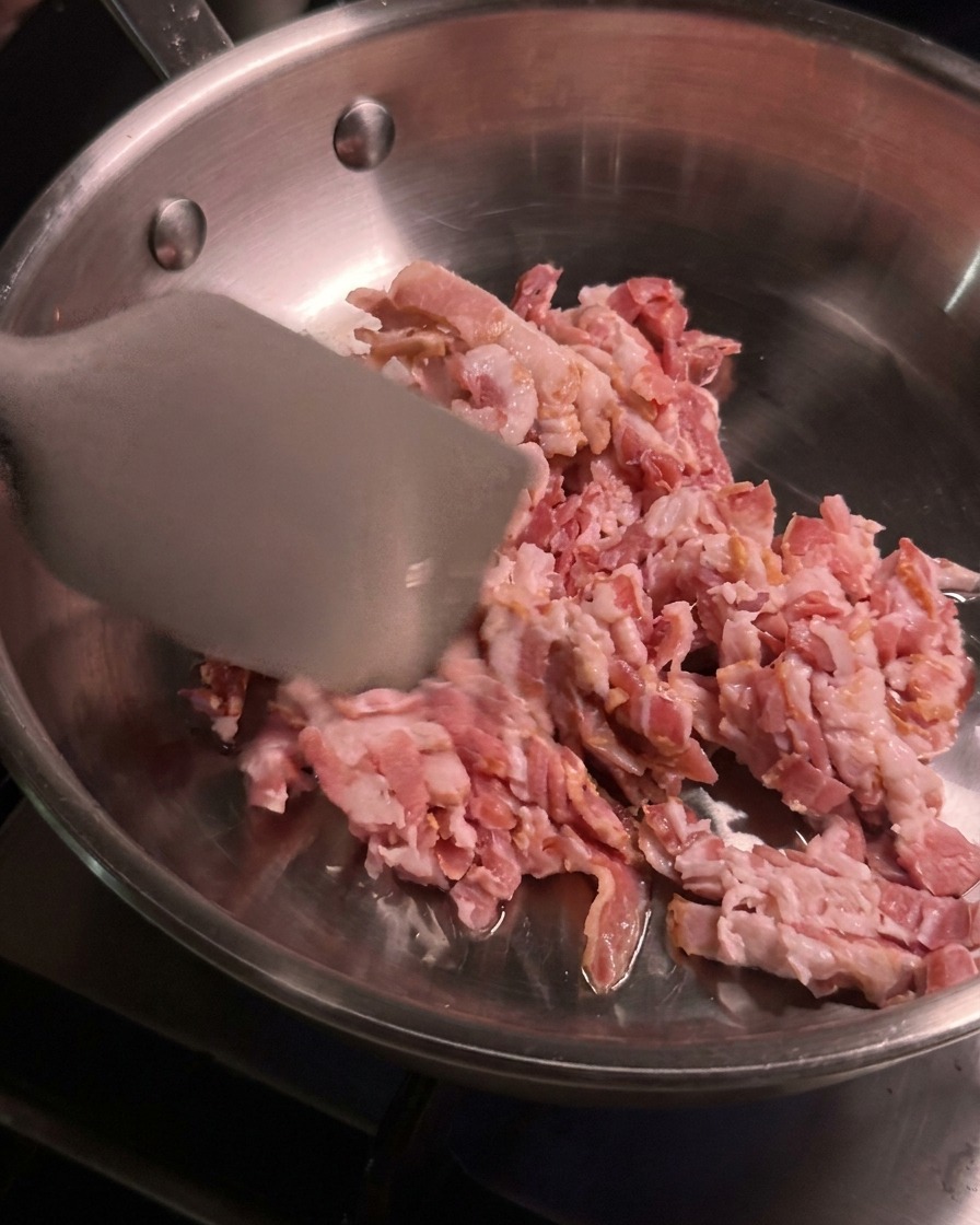 Gewürfelter roher Bacon wird mit einem Pfannenwender aus Metall in einer erhitzten Edelstahlpfanne gewendet.