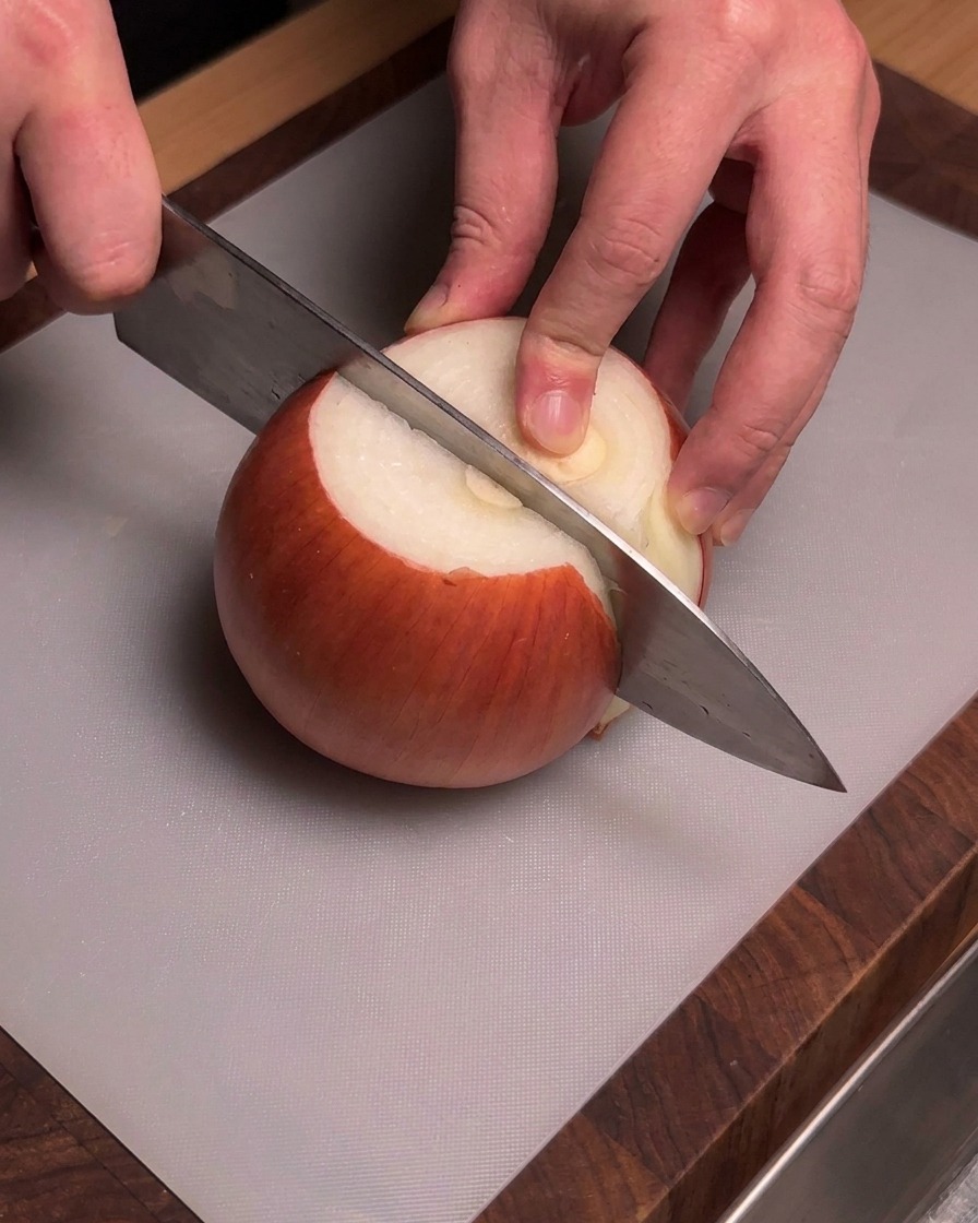 Hände halten ein Kochmesser, um eine geschälte gelbe Zwiebel auf einem grauen Schneidebrett zu schneiden.