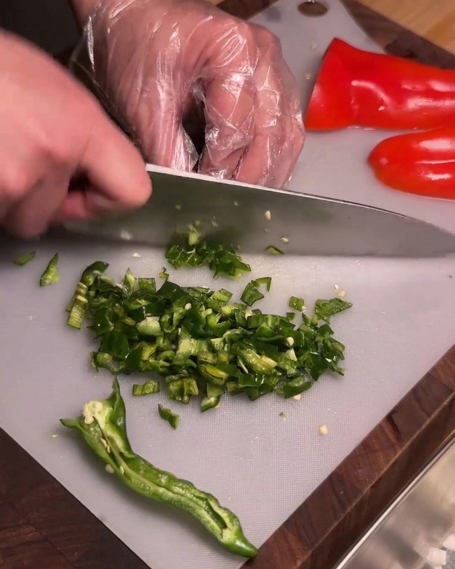 Eine Person mit durchsichtigen Plastikhandschuhen schneidet entkernte grüne Chilis auf einem Schneidebrett in feine Würfel.