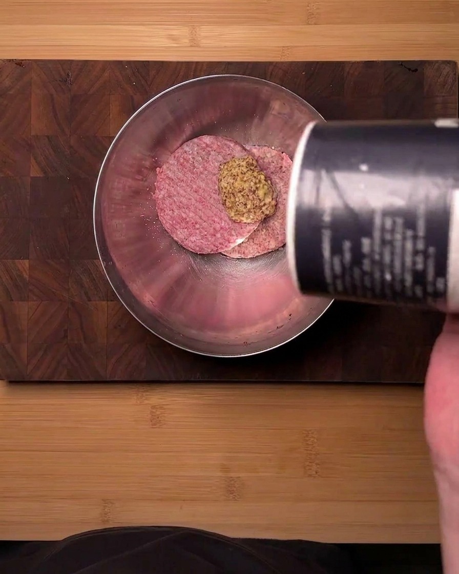 Blick von oben auf rohes Rinderhackfleisch in einer Edelstahlschüssel, das mit Senfpulver und Salz gewürzt wird.