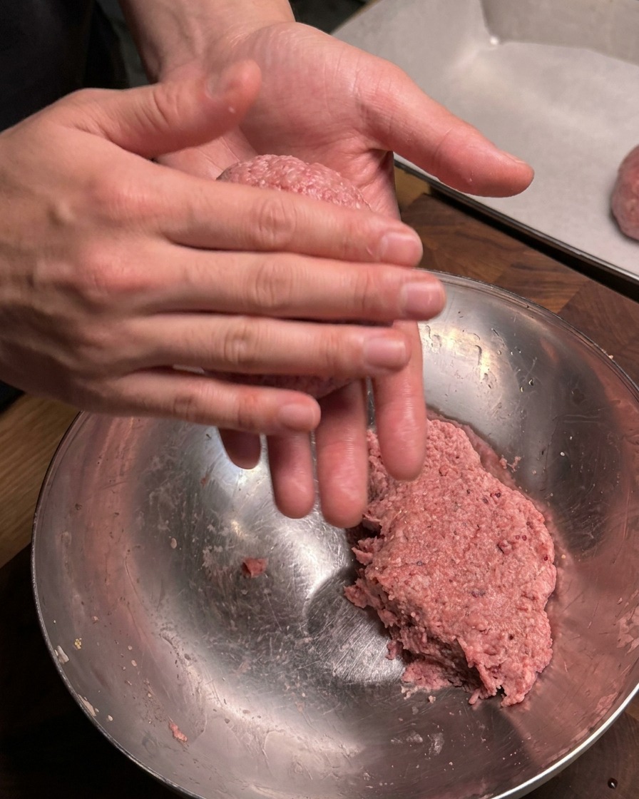 Nahaufnahme von Händen, die fest eine Portion des gewürzten Hackfleischs zu einem runden Fleischbällchen über einer Rührschüssel formen.