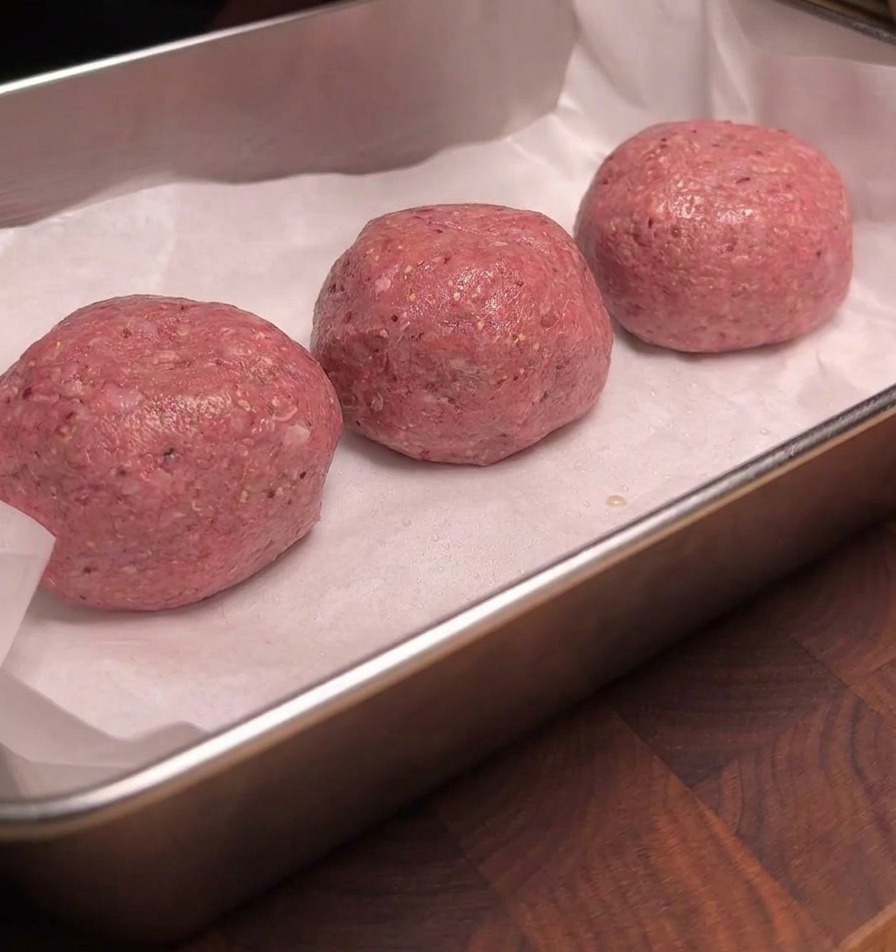 Drei rohe Fleischbällchen liegen auf weißem Backpapier in einem Metallblech, ihre Oberfläche glänzt durch eine Schicht Liquid Smoke.
