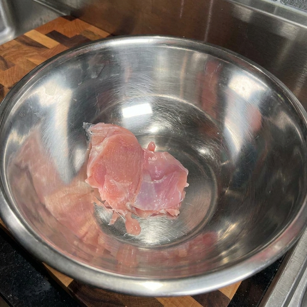 Ein roher Hähnchenschenkel liegt in einer großen, leeren Rührschüssel aus Edelstahl.